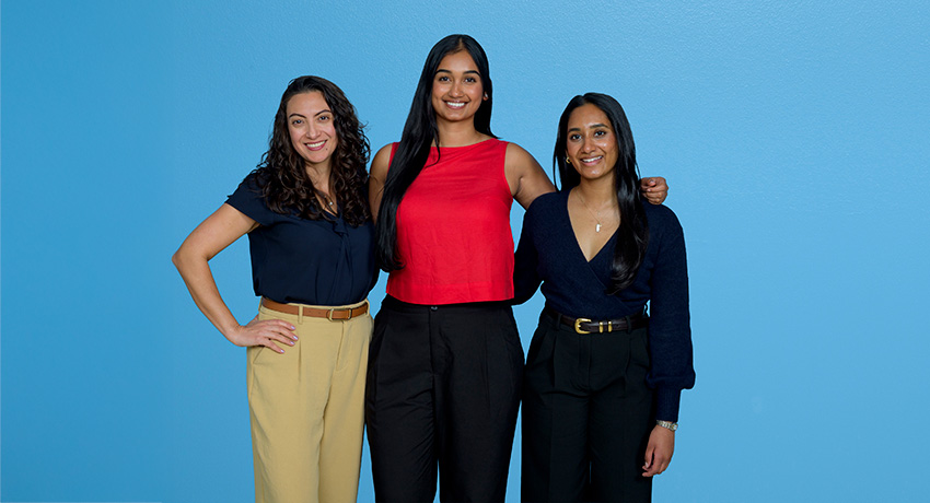 Three women wearing casual business attire stand close together in a group in front of a blue background.