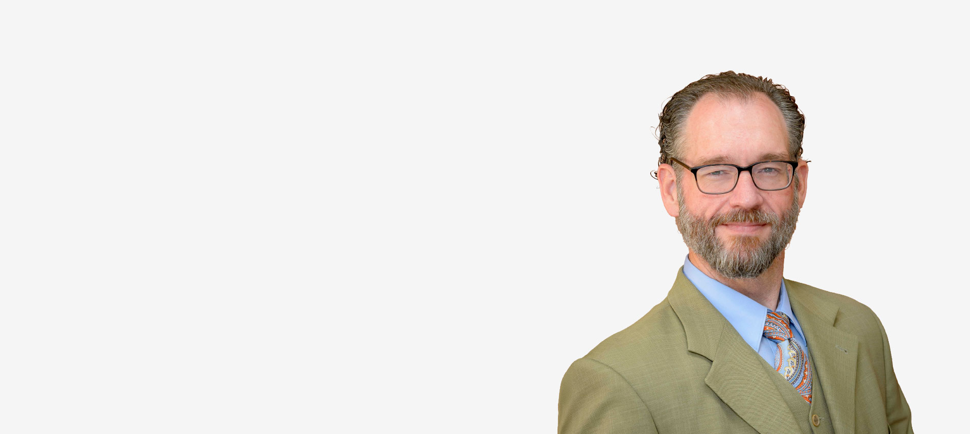 Head-and-shoulders portrait of a person wearing glasses, a light-colored suit jacket, blue shirt, and patterned tie against a plain light gray background. The subject is positioned on the right side of the frame with substantial empty space on the left.