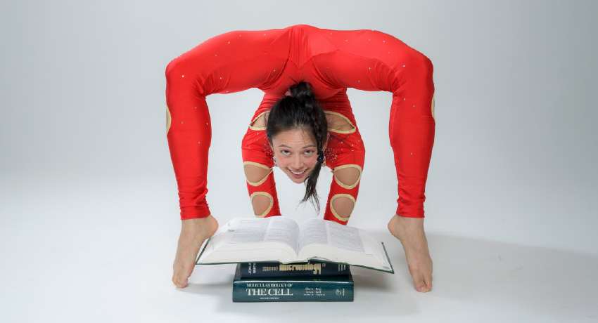 A woman in a red contortion costume balances in an extreme backbend over a stack of medical textbooks with an open book beneath her, smiling toward the camera against a white background.