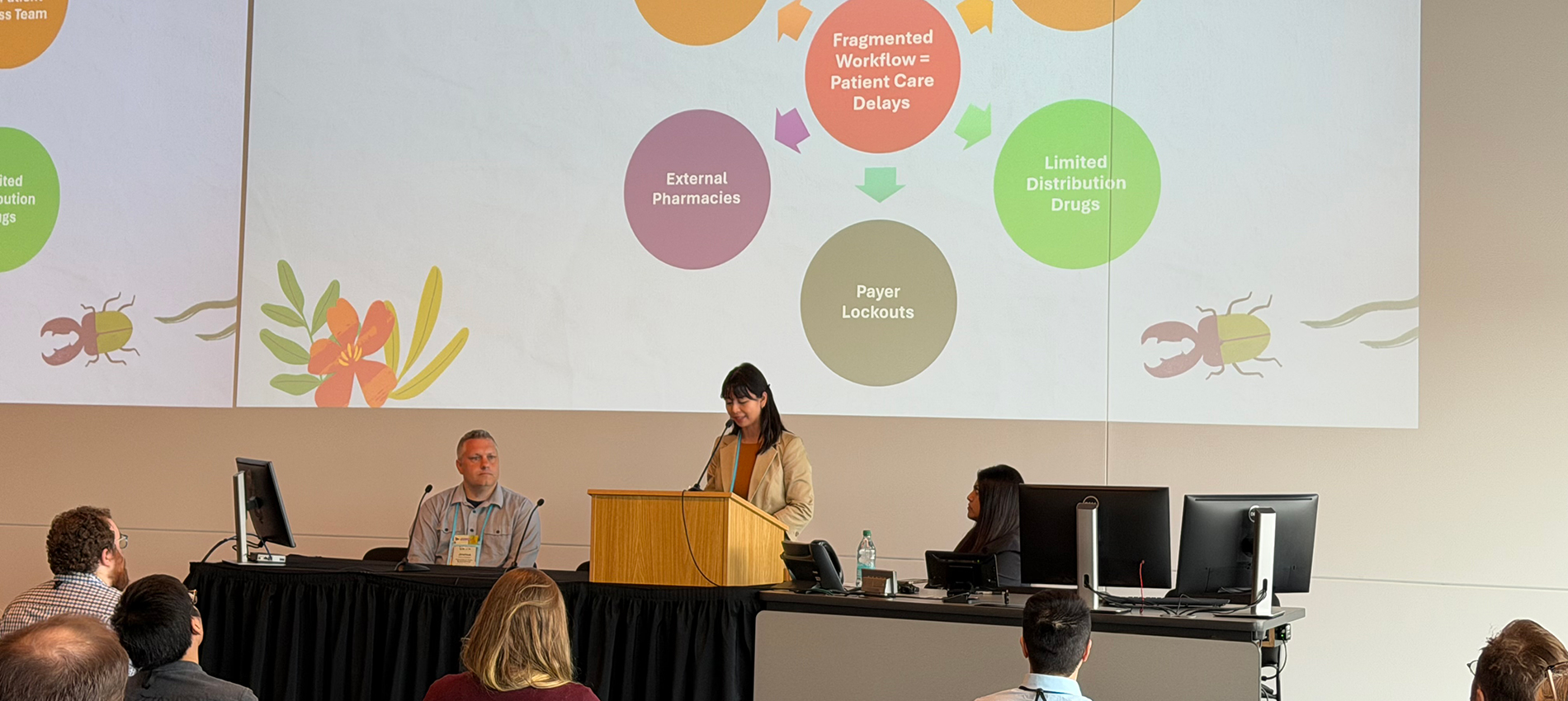 A woman stands at a podium with a very large slide presentation projected behind her. Two co-presenters look on.