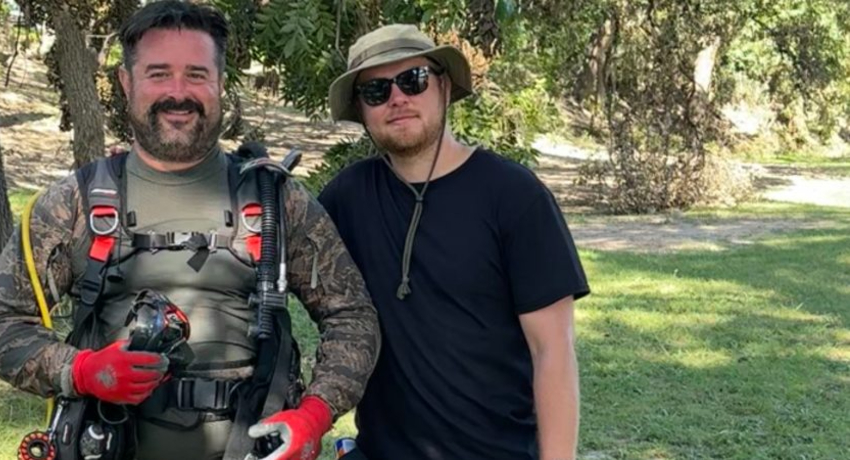 Two people standing outdoors on grass, one wearing protective diving gear with a tank, regulator, and red gloves, and the other wearing a dark shirt, wide‑brim hat, and sunglasses. Trees and a sunlit path are visible in the background.
