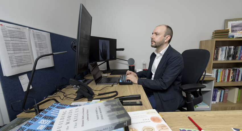 Jacob Moses, PhD, assistant professor in the Department of Bioethics and Health Humanities at UTMB, sits at his office desk working on a computer. Books, research materials and multiple monitors surround his workspace as he studies healthcare ethics.