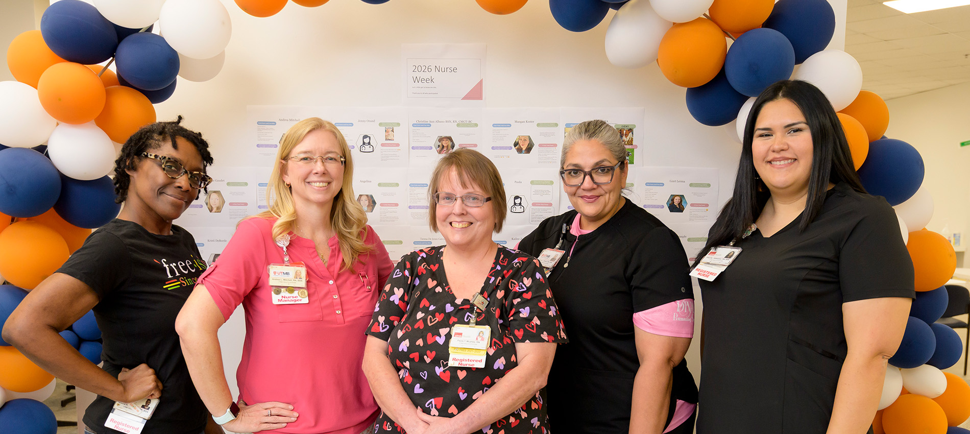 Five nurses stand together smiling beneath a blue, orange, and white balloon arch during a 2026 Nurse Week celebration. A display board featuring staff profiles is visible behind the group as they pose in a hospital or clinic setting.