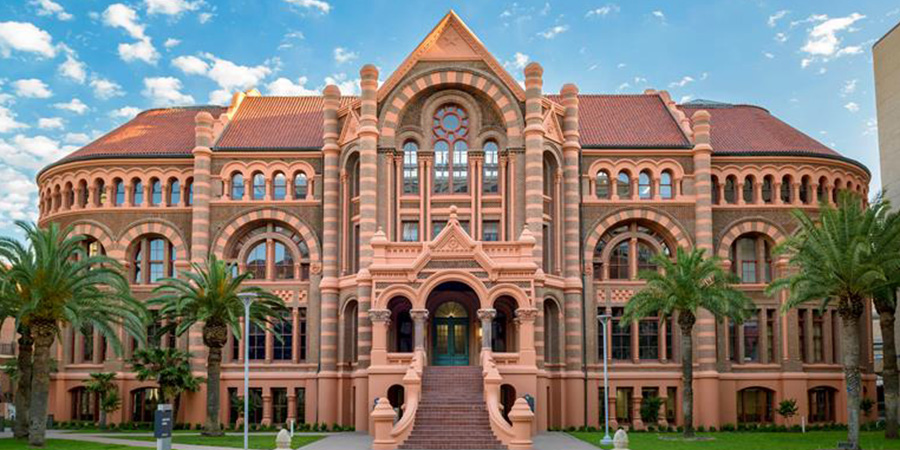 Exterior view of UTMB’s historic campus building with red‑brick Romanesque architecture, arched windows, palm trees, and a central staircase under a partly cloudy sky