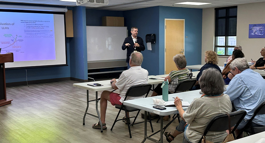 A man in a suit stands in front of seated students over 50 who want to learn about AI.