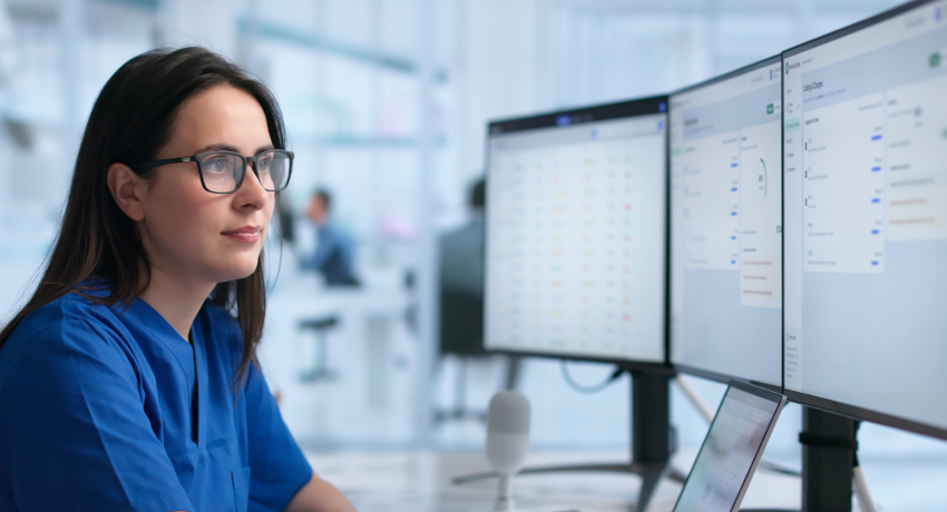 A woman in blue scrubs is seated at a workstation, looking at two computer monitors displaying software interfaces. A laptop is open on the desk in a clinical or office setting.