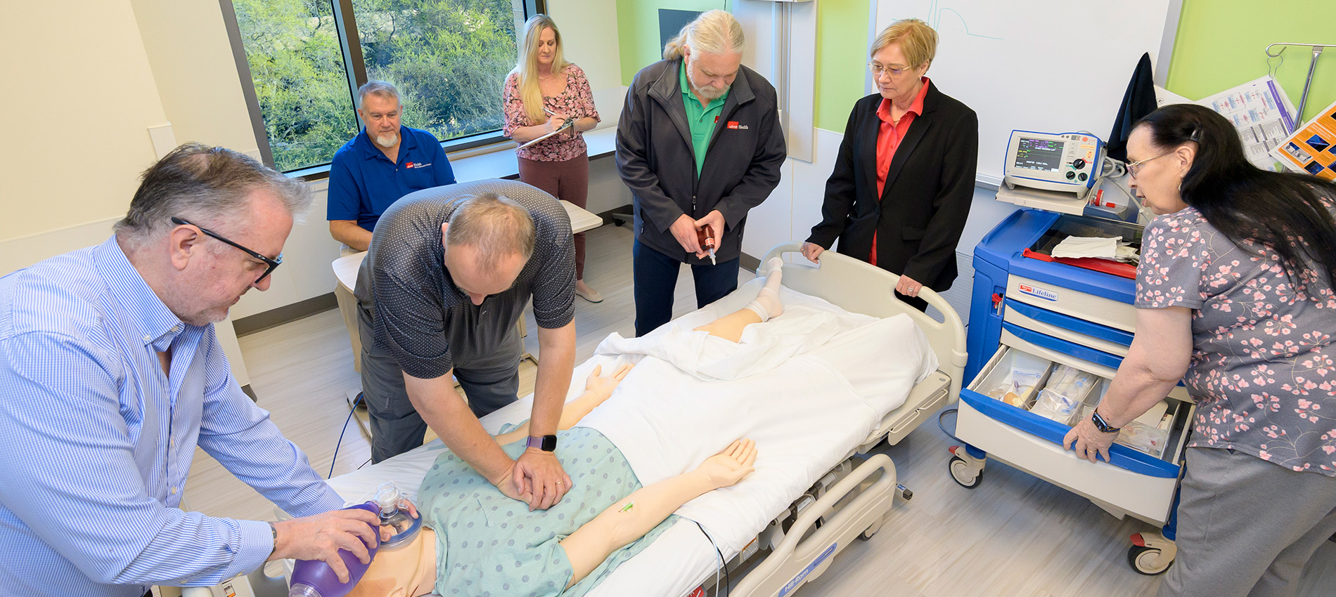 Two instructors demonstrate proper CPR technique. One executes chest compressions, while the other holds a mask on the CPR dummy's face. Five other instructors observe the action.