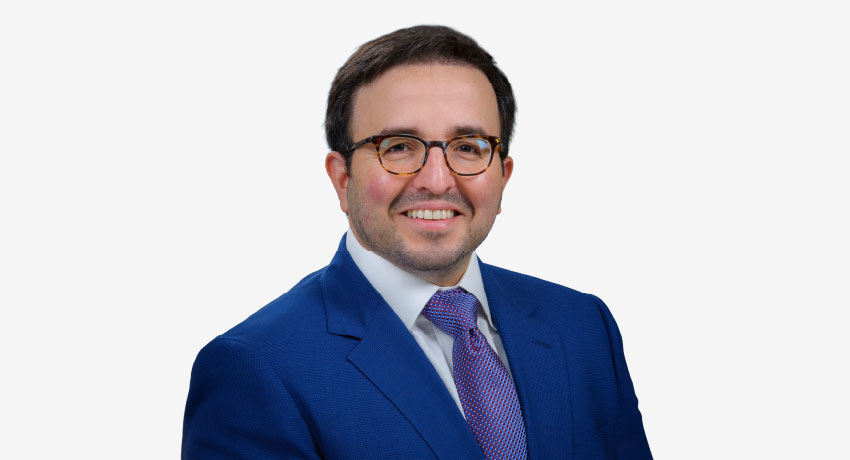 Professional head-and-shoulders portrait shows a man wearing glasses, a blue suit jacket, white shirt, and patterned tie against a light gray background.