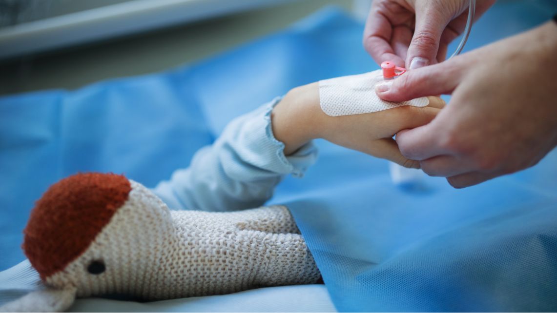 Close‑up of a health care worker placing a small bandage and tubing on a child’s arm resting on a blue medical drape