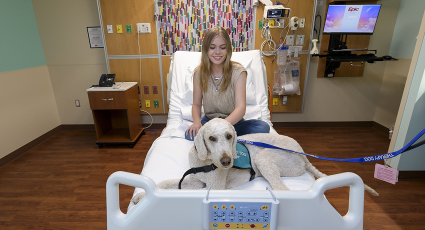 A teenage patient smiles from her hospital bed as a therapy dog wearing a blue service vest rests beside her. The dog provides comfort and emotional support during her hospital stay, helping create a calmer, more reassuring environment.