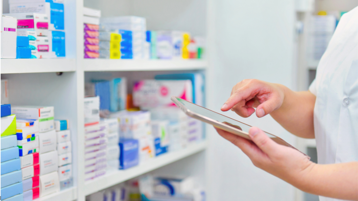 Hands holding a tablet in a pharmacy storage area, with shelves of medication boxes in the background, representing hands-on clinical training for pharmacy students at UTMB