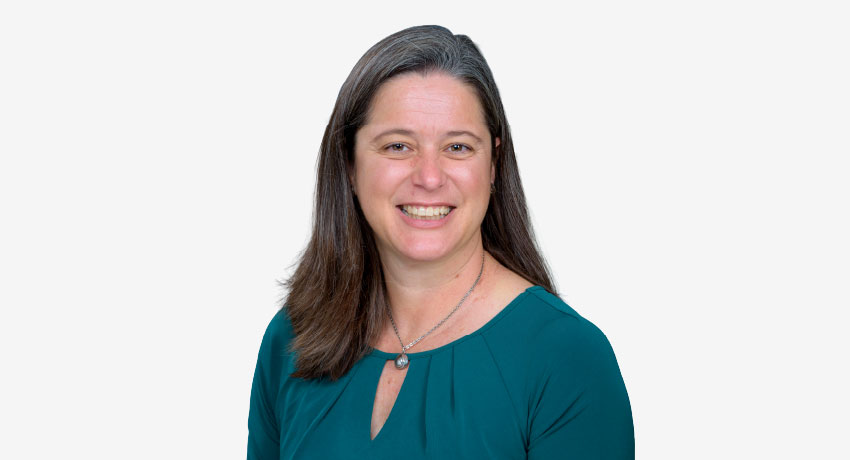 Formal head-and-shoulders studio portrait of a woman with long brown hair wearing a teal top and a necklace against a white background.