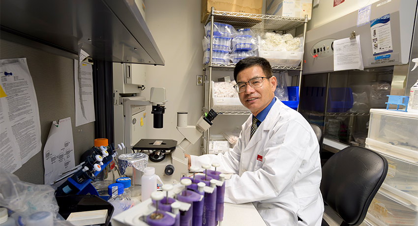 A man in a white lab coat sits at a laboratory workstation beside a microscope, smiling toward the camera. Shelves stocked with medical and laboratory supplies are visible in the background, while rows of capped sample tubes sit in the foreground.