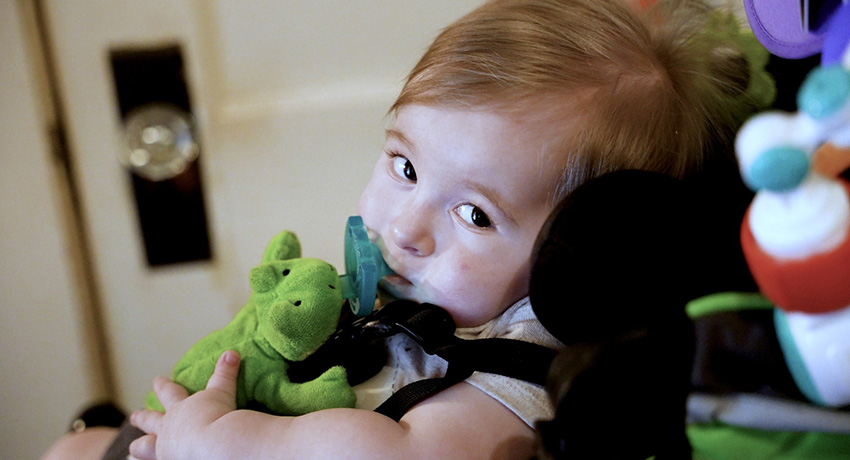 Baby holding a green stuffed toy while seated indoors near colorful toys.