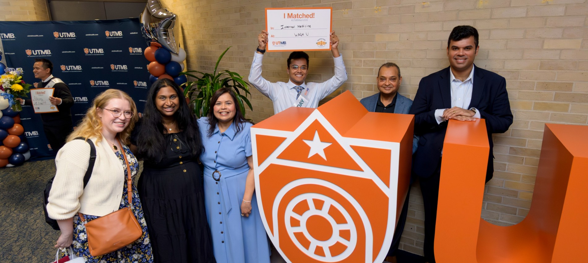 Group of six smiling people pose at a UTMB Match Day celebration, standing beside large orange “UTMB” letters and a shield logo, while one person in the center holds up a “I Matched!” sign