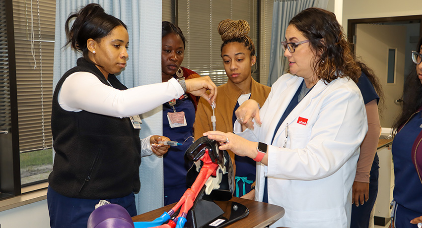 An instructor in a white coat instructs a respiratory therapy student who is inserting a tube into the mouth of a model of a human airway.