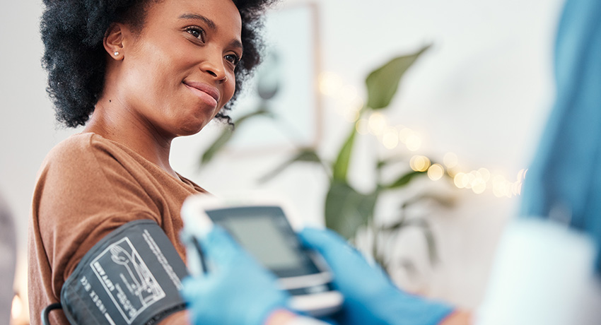 A person seated with a blood pressure cuff on the upper arm while a gloved clinician holds a digital blood pressure monitor; the person’s face is intentionally blurred.