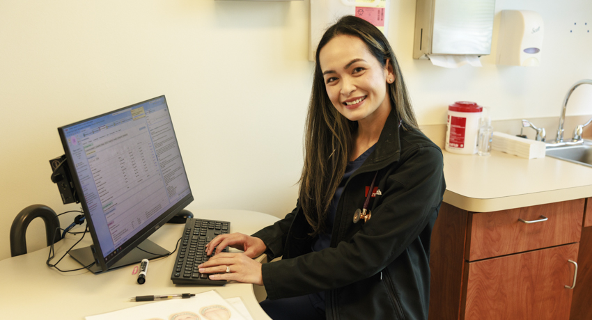 UTMB nurse practitioner Ruth Diamampo Jetty sits at a computer in a clinical exam room, smiling at the camera. She wears a dark jacket over scrubs and has a stethoscope around her neck.