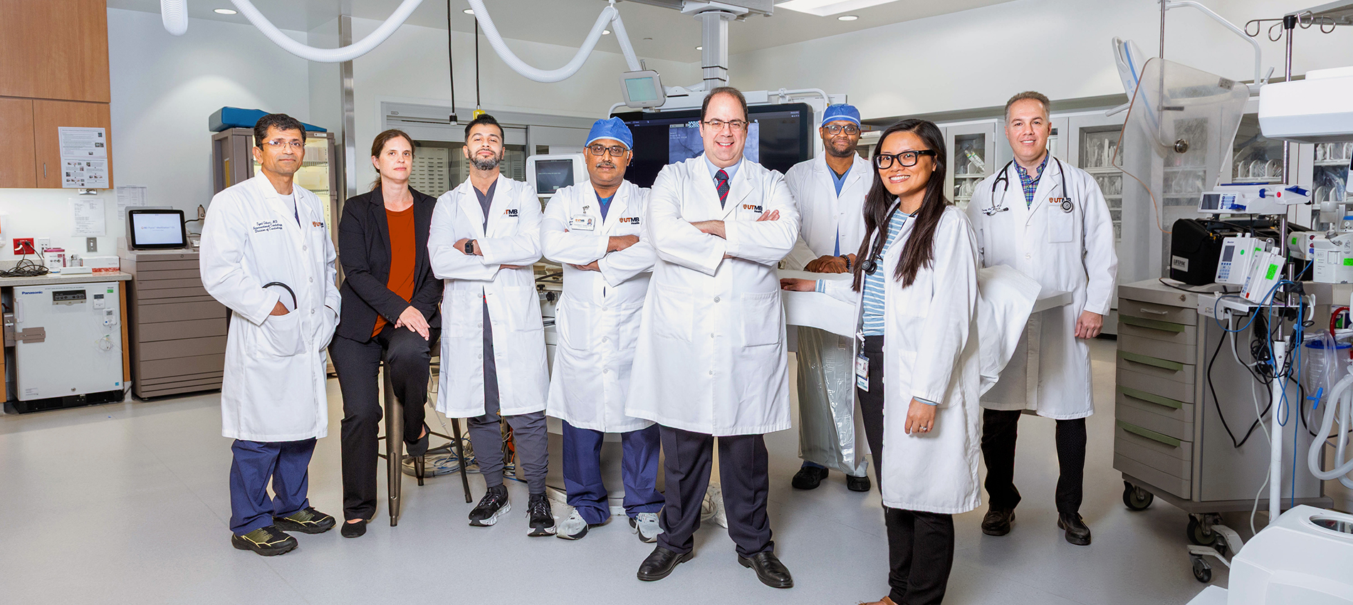 Seven men and women in UTMB white coats and one woman in black business attire stand in a procedure room.
