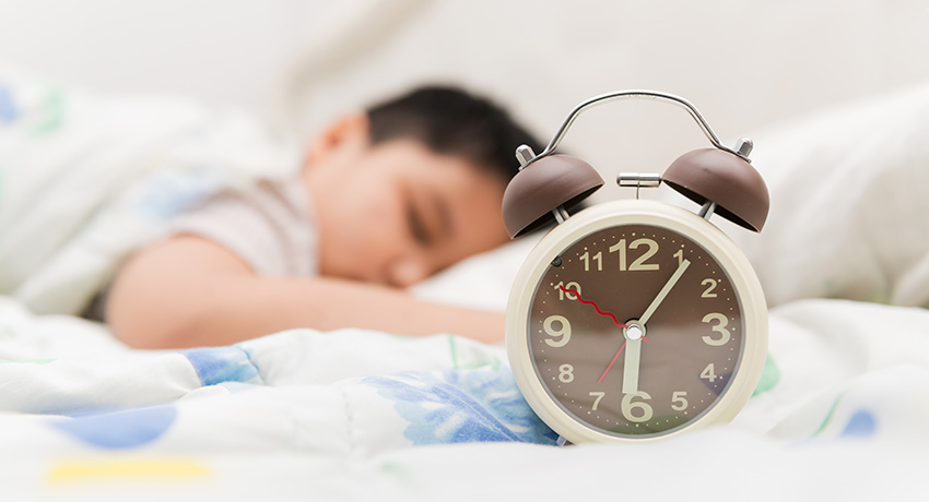 Alarm clock on a bed in the foreground with a child sleeping out of focus in the background.