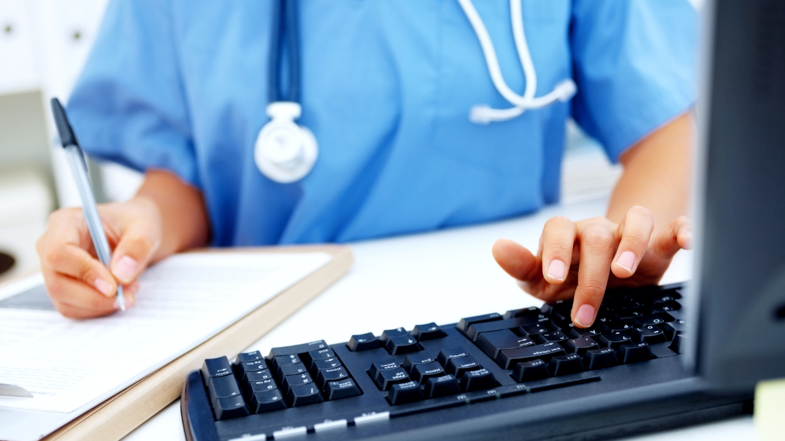 A nurse in blue scrubs works at a desk with a computer and a clipboard