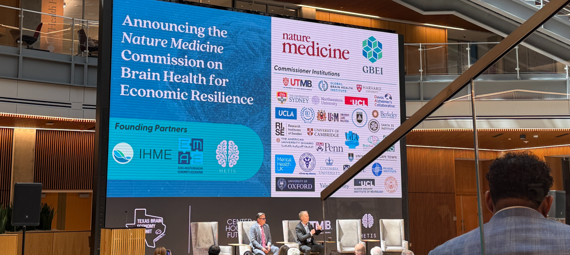 Large conference screen displays “Announcing the Nature Medicine Commission on Brain Health for Economic Resilience,” alongside logos of UTMB and other institutions; two speakers sit on a stage below with chairs and audience visible.