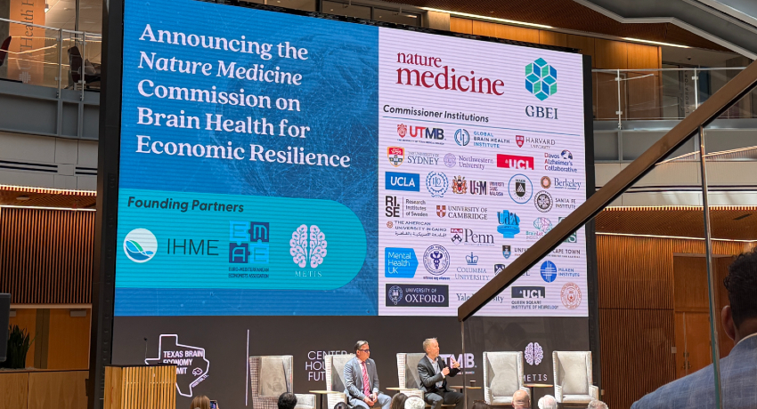 Large conference screen displays “Announcing the Nature Medicine Commission on Brain Health for Economic Resilience,” alongside logos of UTMB and other institutions; two speakers sit on a stage below with chairs and audience visible.