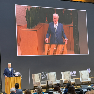 Texas Lt. Gov. Dan Patrick addresses attendees at the Texas Brain Economy Summit. A speaker stands at a podium addressing an audience while a large screen above shows a live close-up of the same speaker; conference branding and empty stage chairs appear in the background.