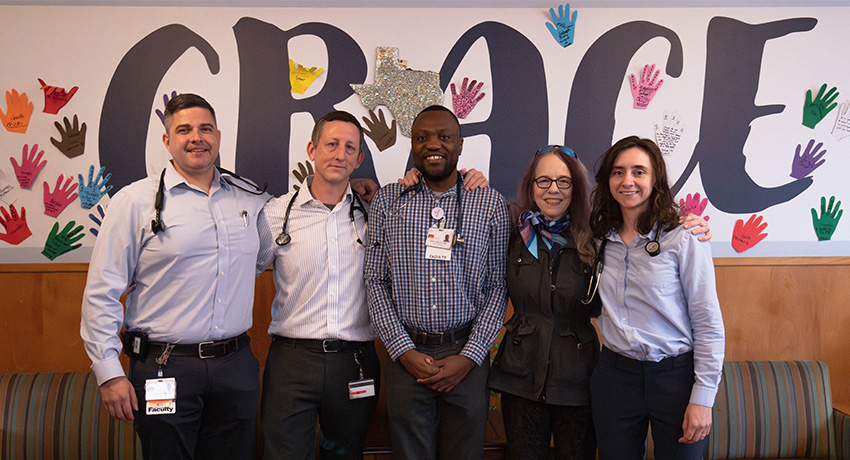Five staff members wearing ID badges and stethoscopes stand side by side in front of a wall display reading “GRACE,” decorated with colorful handprints and a Texas-shaped element.