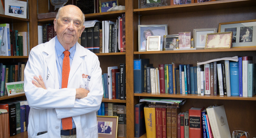 Man in a white lab coat and orange tie is shown standing with arms crossed in front of bookshelves filled with books and framed photographs in an office.