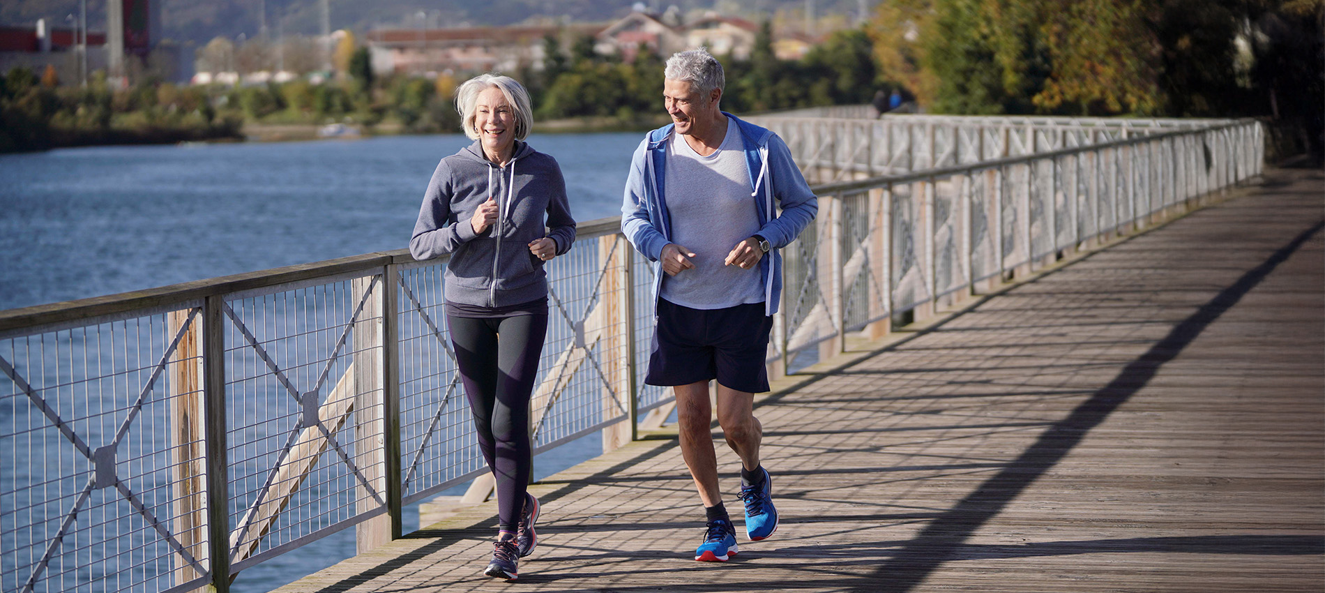 Two senior citizens run on a bridge across a body of water.