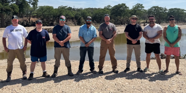 Tyler Morrison and Austin Johnson worked alongside the United Cajun Navy in the aftermath of the flooding in Kerr County. Eight people stand in a line on a rocky riverbank with water and trees in the background. Most wear caps or sunglasses and casual outdoor clothing, with hands clasped in front.