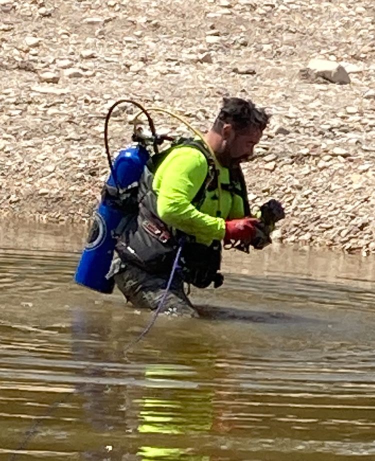 Person in a high-visibility shirt and flotation vest with an air tank wades through shallow water along a rocky bank while handling a small dark object.