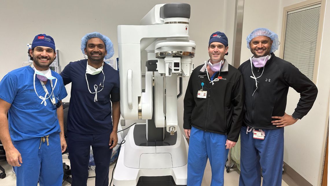 Four UTMB urology team members wearing blue surgical scrubs and caps stand in an operating room beside a single‑port robotic surgery system
