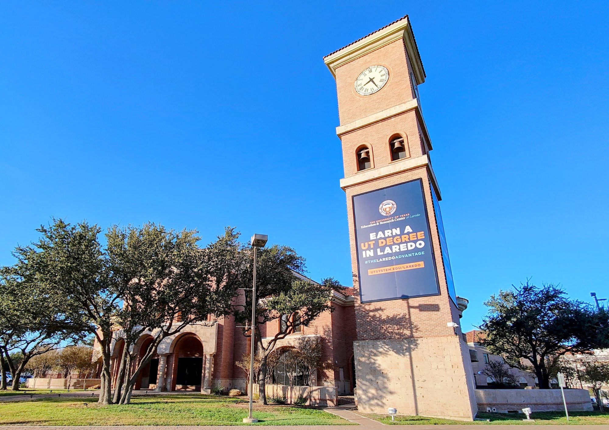 Exterior view of the UT Education and Research Center at Laredo, featuring a brick building with a tall clock tower and a banner reading “Earn a UT Degree in Laredo” under a clear blue sky