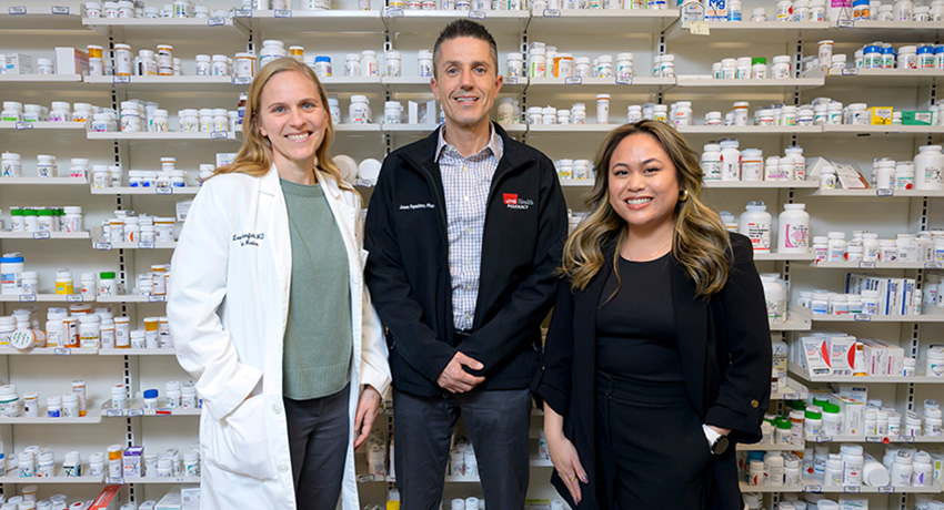 Three pharmacy team members standing in front of shelves filled with prescription medication bottles.