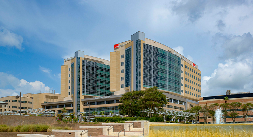 Tiered water fountain in the foreground with a large modern hospital building rising behind it under a partly cloudy sky.