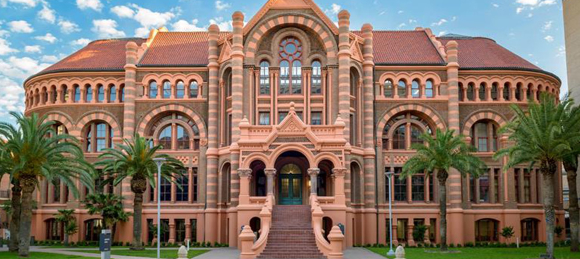 Exterior view of UTMB’s historic campus building with red‑brick Romanesque architecture, arched windows, palm trees, and a central staircase under a partly cloudy sky