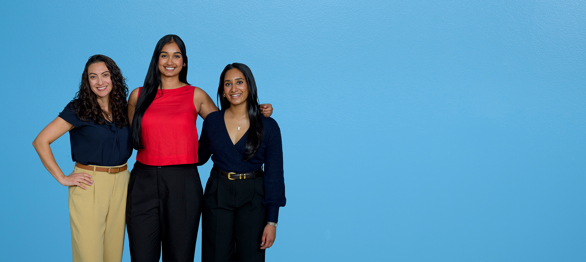 Three women wearing casual business attire stand close together in a group in front of a blue background.