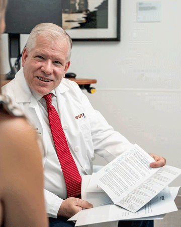 UTMB orthopedic surgeon Dr. Paul Dicpinigaitis. Person in a white medical coat and red tie sits holding several printed documents while speaking with another seated person in an office setting.