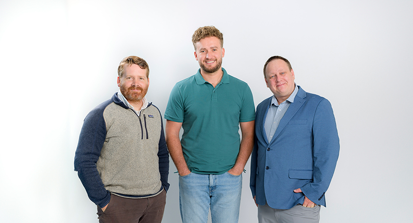 Three men in business casual clothing stand together agains a white background.