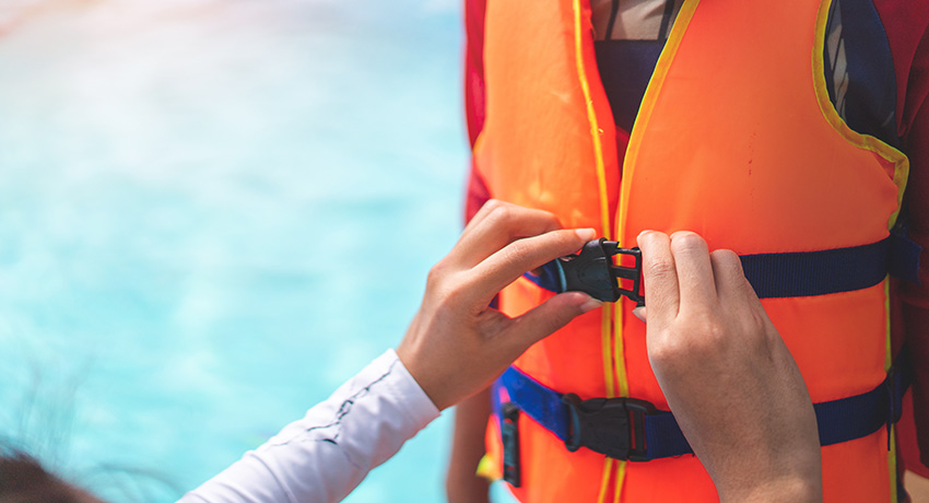 Person fastening the front buckle of an orange life jacket while standing beside water.