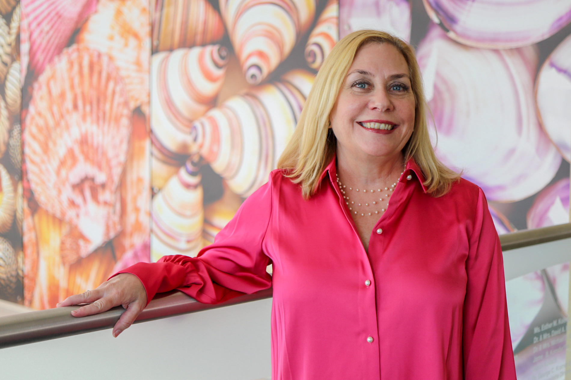 An individual standing indoors against a colorful seashell-themed wall, wearing a bright button-up top and resting one hand on a railing.
