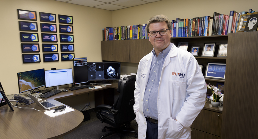 Dr. Max Wintermark, wearing a white coat bearing the UTMB logo, stands near his desk.