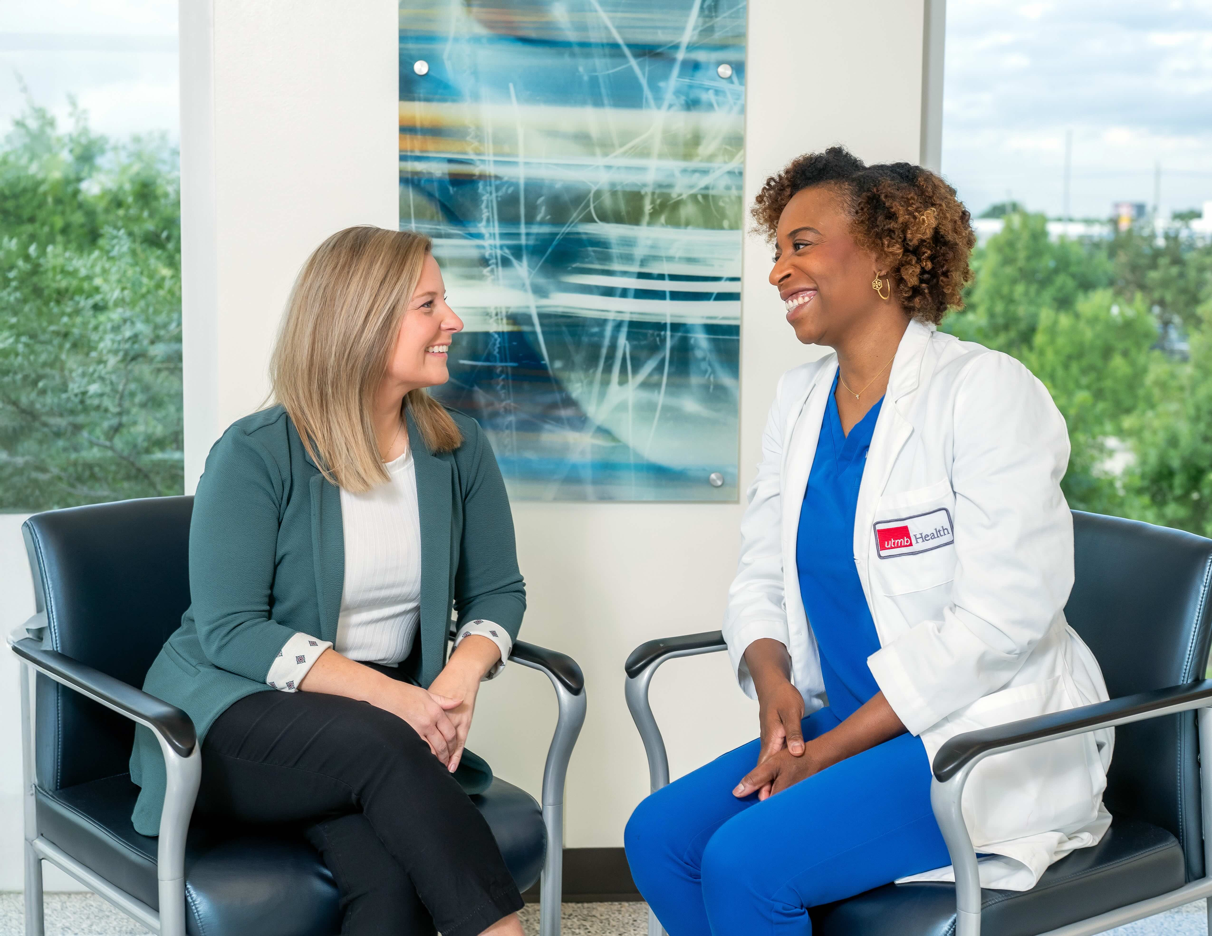 A patient and a health care provider sit facing each other in chairs during an office visit, with large windows and framed artwork visible in the background