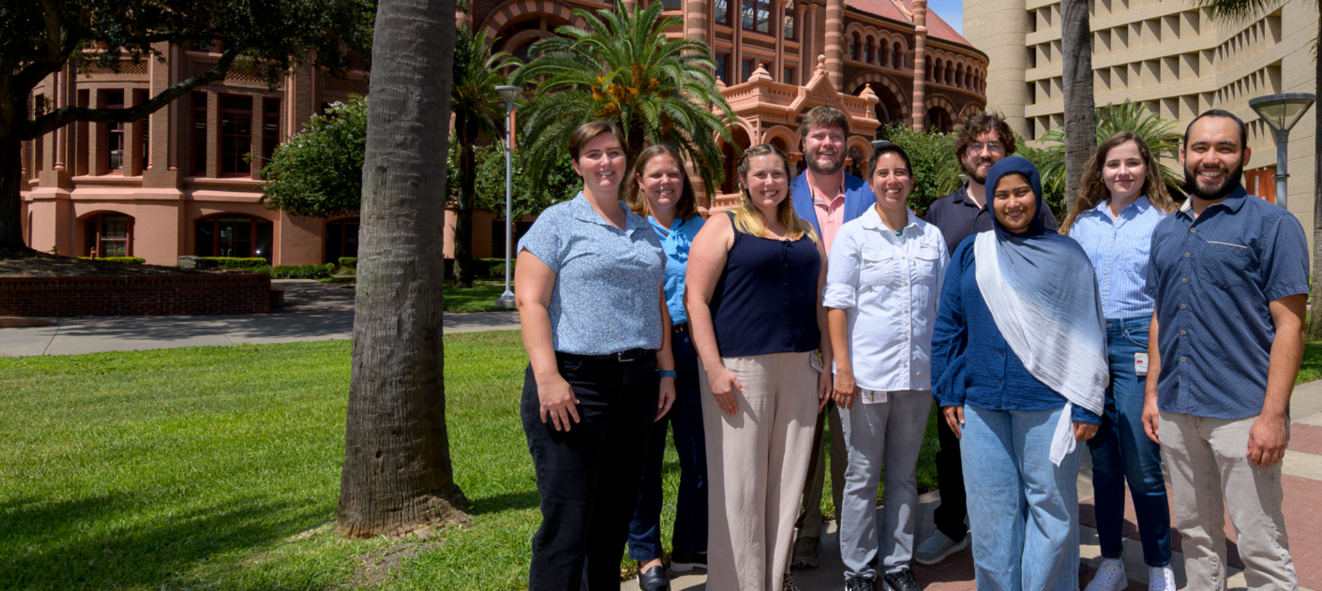 A group of eight people is standing outdoors on a lawn in front of a historic red-brick building and adjacent campus buildings, with palm trees visible in the background.