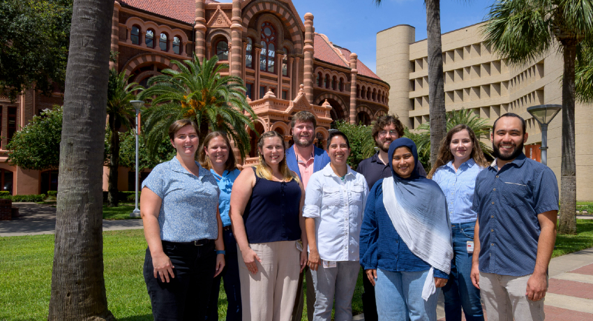 A group of eight people is standing outdoors on a lawn in front of a historic red-brick building and adjacent campus buildings, with palm trees visible in the background.