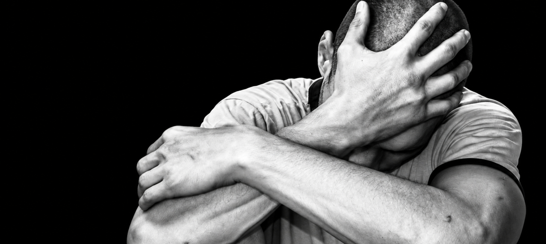 A man is shown seated with knees drawn up, one arm wrapped around his legs and a hand covering part of his head against a dark background.