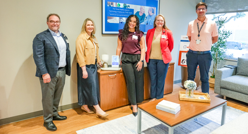 Five people stand together in an office setting near a cabinet and coffee table. A wall-mounted display screen and seating area are visible in the background.