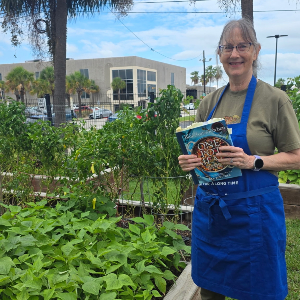 H A person wearing a blue apron stands in a raised garden bed area and holds a cookbook. Garden plants, trees, and a building are visible in the background.  